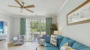 Sala de estar com ventilador de teto, sofá, duas poltronas, mesa de centro e vista para uma área externa com um alpendre.