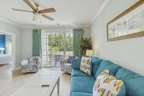 Ein Wohnzimmer mit Deckenventilator, einer Couch, zwei Sesseln, einem Couchtisch und Blick durch eine Glastür auf einen Außenbereich mit Terrasse.