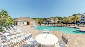Uma área ao lado da piscina com espreguiçadeiras e uma mesa, cercada por edifícios e palmeiras.