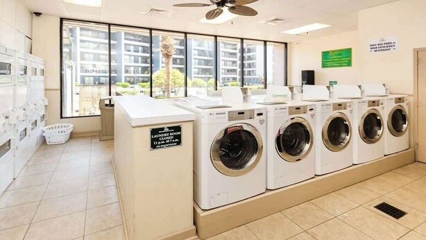 A laundry room with a row of washing machines, a sign indicating a 24-hour service, and a view of the outside through large windows. - Renaissance Tower 317 (Myrtle Beach)
