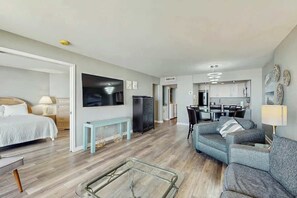 A modern living room with a flat-screen TV, a sofa, a glass coffee table, and a dining area with a kitchen in the background. - Ocean Creek Unit G-8 (Myrtle Beach)