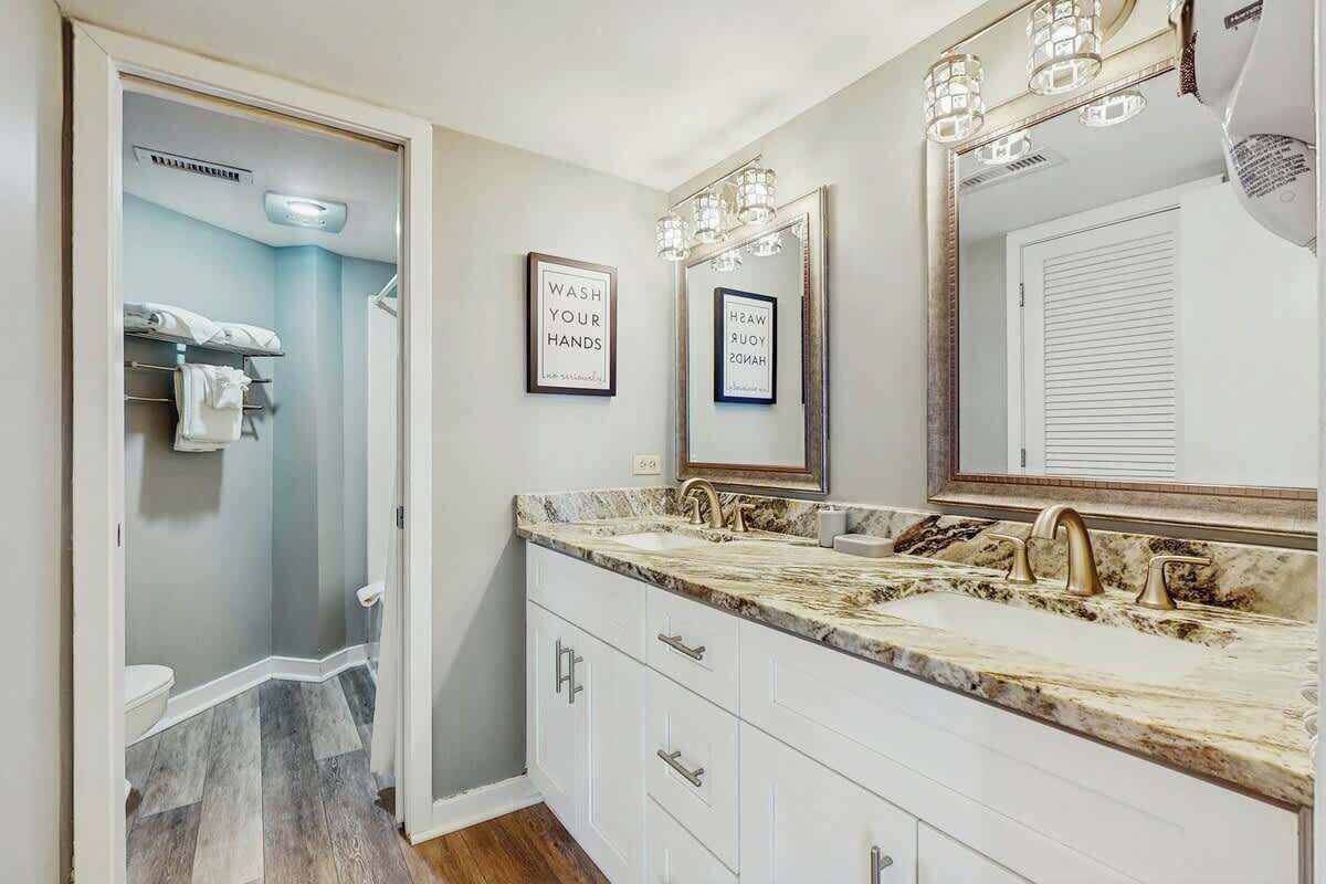 A bathroom with a double sink, vanity, and a mirror with lights. There are framed signs on the wall and a towel rack.