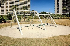 A swing set in a sandy area with a building in the background. - Ocean Creek Unit G-8 (Myrtle Beach)