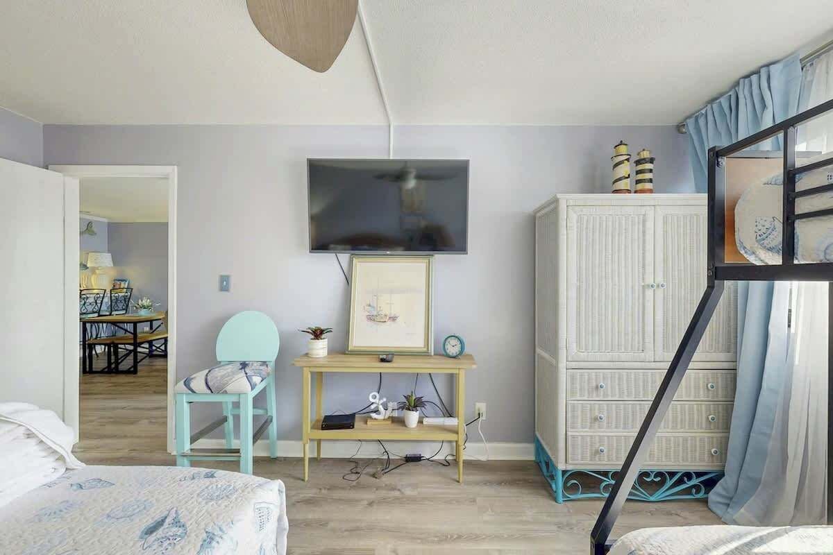 A bedroom with a bed, a TV mounted on the wall, a small table with a chair, and a wardrobe.