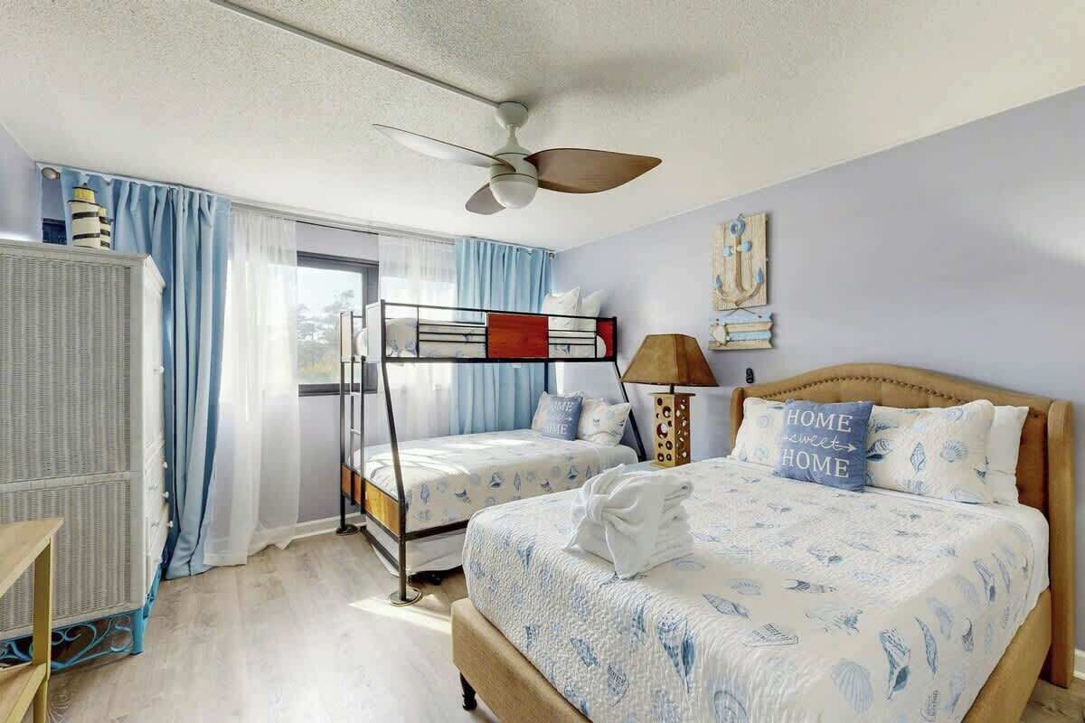 Ein Hotelzimmer mit Etagenbett, einem Einzelbett, einem Deckenventilator, einer Heizung und einem Fenster mit Vorhängen.