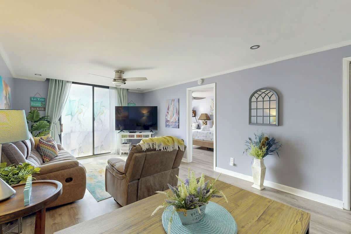 A modern living room with a sofa, a dining table with a plant, a television, and a ceiling fan.
