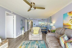 A living room with a ceiling fan, a sofa, a chair, a rug, and a dining area in the background. - Holiday Towers Unit 301 (Myrtle Beach)
