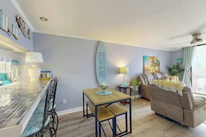 A living room with a surfboard decoration, a sofa, a dining table, and a bar area. - Holiday Towers Unit 301 (Myrtle Beach)