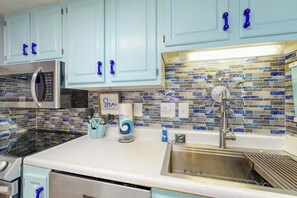 A kitchen with light blue cabinets, a stainless steel sink, and a tiled backsplash. - Holiday Towers Unit 301 (Myrtle Beach)