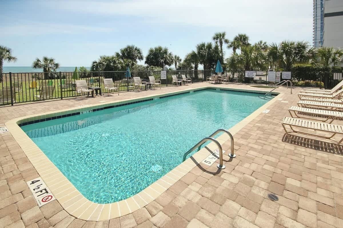 A rectangular swimming pool with a tiled edge, surrounded by lounge chairs and a fence.