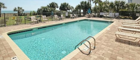 Une piscine rectangulaire dont le bord est recouvert de carreaux, entourée de chaises longues et d’une clôture.