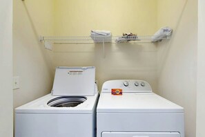 A laundry room with two white washing machines and a metal shelf above them. - Myrtlewood Golf & Beach Retreat (Myrtle Beach)