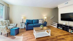 A living room with a blue sofa, a patterned armchair, a coffee table, and a TV on a stand. - Brandywine South Unit 4G (Myrtle Beach)
