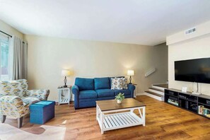 Ein Wohnzimmer mit blauem Sofa, einem gemusterten Sessel, einem Couchtisch und einem Fernseher auf einem Schrank.