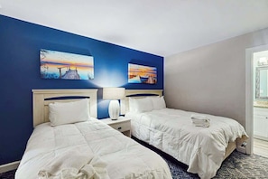 A hotel room with two beds, a nightstand with a lamp, and two paintings on the wall. - Chelsea House Villas 112 (Myrtle Beach)