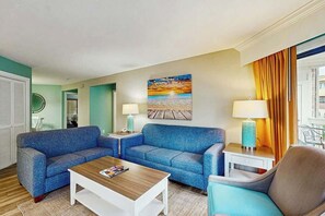 A hotel room with a blue sofa set, a wooden coffee table, a painting of a sunset, and a view of the ocean through the window. - Chelsea House Villas 112 (Myrtle Beach)