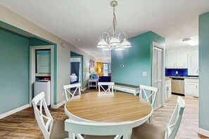A dining area with a round table and white chairs, a kitchen in the background, and teal walls. - Chelsea House Villas 112 (Myrtle Beach)