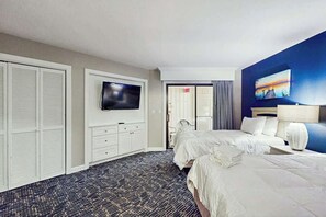 A hotel room with two beds, a flat-screen TV mounted on the wall, a desk with a lamp, and a painting on the wall. - Chelsea House Villas 112 (Myrtle Beach)