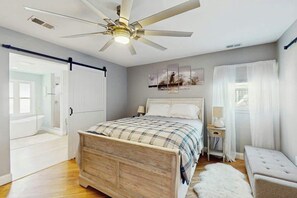 A bedroom with a bed, a bedside table, a lamp, a ceiling fan, and a window with curtains. - Beach House Hideaway (North Myrtle Beach)