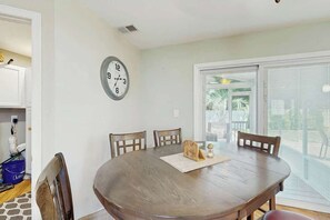 A dining area with a round table, four chairs, a clock on the wall, and a sliding glass door leading outside. - Beach House Hideaway (North Myrtle Beach)