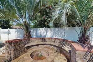 A curved stone fire pit with a brick border, surrounded by palm trees and a white fence. - Beach House Hideaway (North Myrtle Beach)