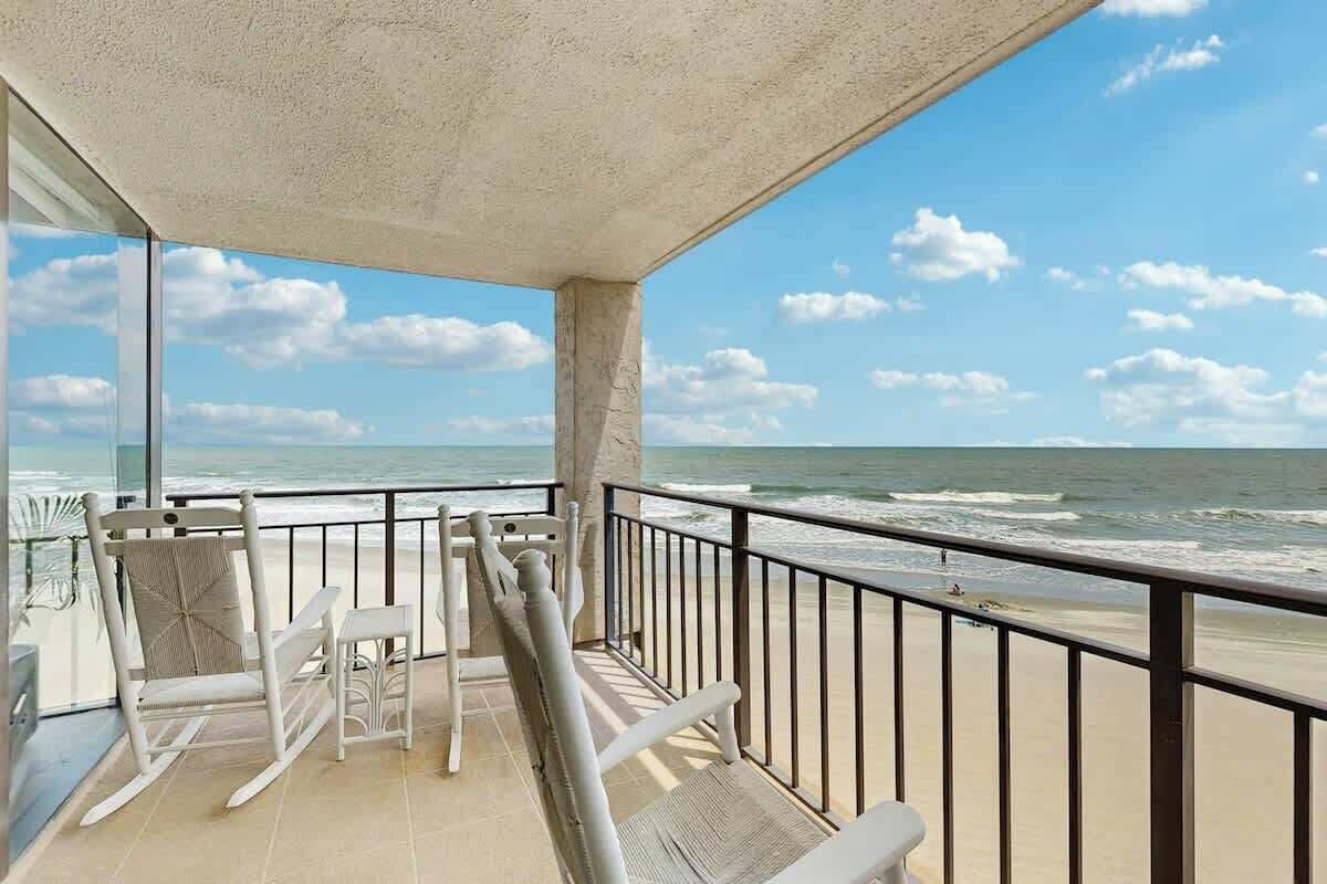 A balcony with a view of the ocean, a white rocking chair, a small table, and a glass door leading to the beach.