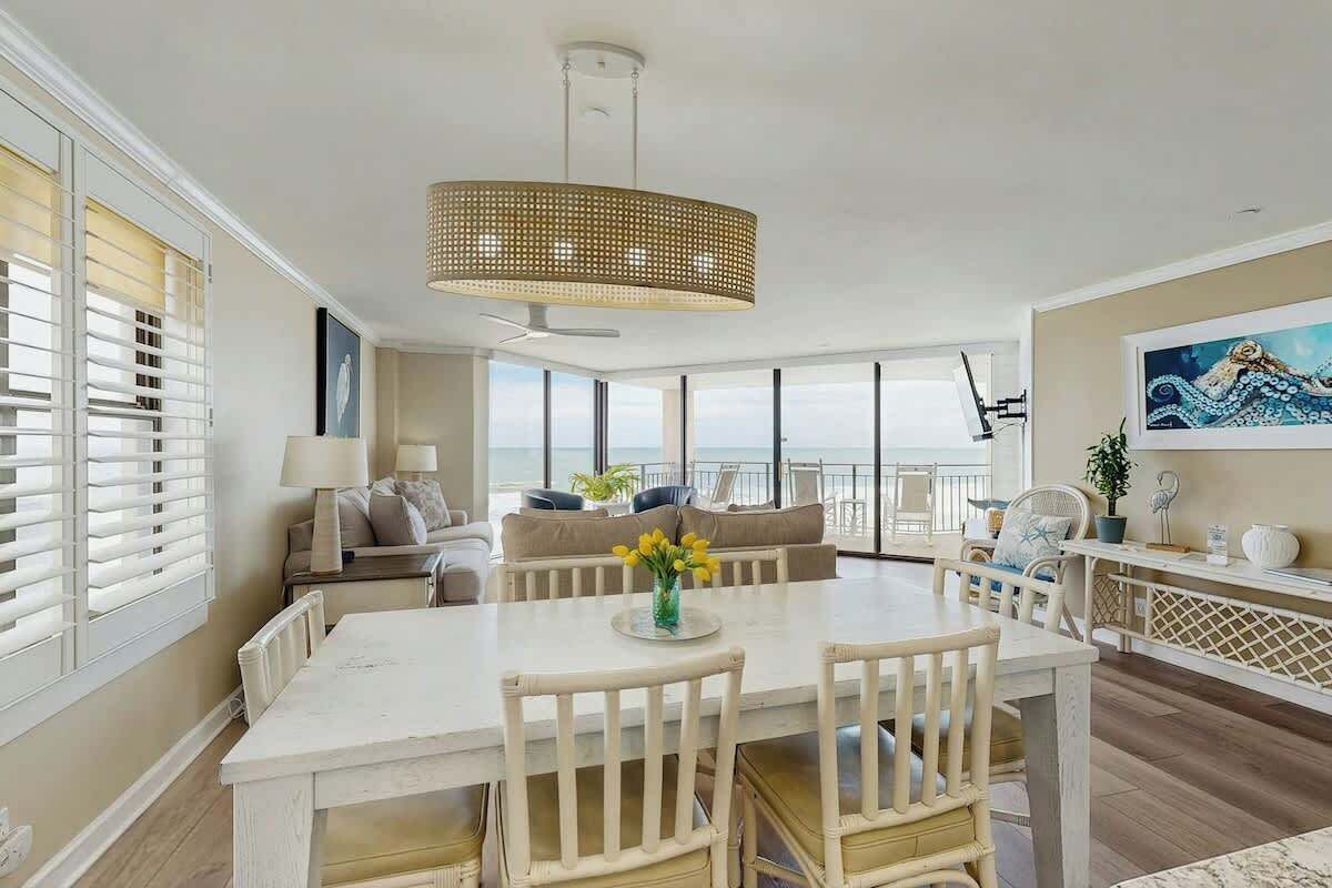 A spacious dining area with a white table and chairs, a hanging light fixture, and a view of the ocean through large windows.