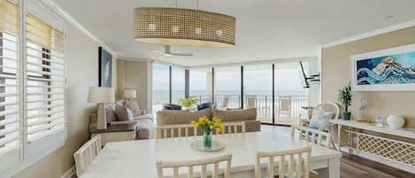 Uma sala de jantar espaçosa com uma mesa e cadeiras brancas, um lustre pendurado e vista para o oceano através de amplas janelas.