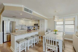 A modern kitchen with white cabinetry, a central island with stools, and a dining area with a table and chairs. - Surfmaster by the Sea 208 (Murrells Inlet)