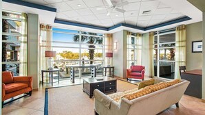 A spacious living area with a sofa, a red armchair, a coffee table, and a TV. There are bookshelves on the left and a large window with a view of palm trees. - Harbourgate Unit 0620 (North Myrtle Beach)
