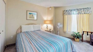 Une chambre d’hôtel comprenant un lit, une table de chevet, une lampe, un ventilateur, une fenêtre avec des rideaux et une petite plante.