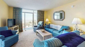 Une chambre d’hôtel avec un canapé, un fauteuil, une télévision et un balcon doté de rideaux.