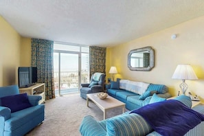 Ein Hotelzimmer mit Sofa, Sessel, Fernseher und einem Balkon mit Vorhängen.