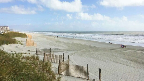 Une plage avec une clôture en bois, un sentier sablonneux et quelques personnes qui se promènent.