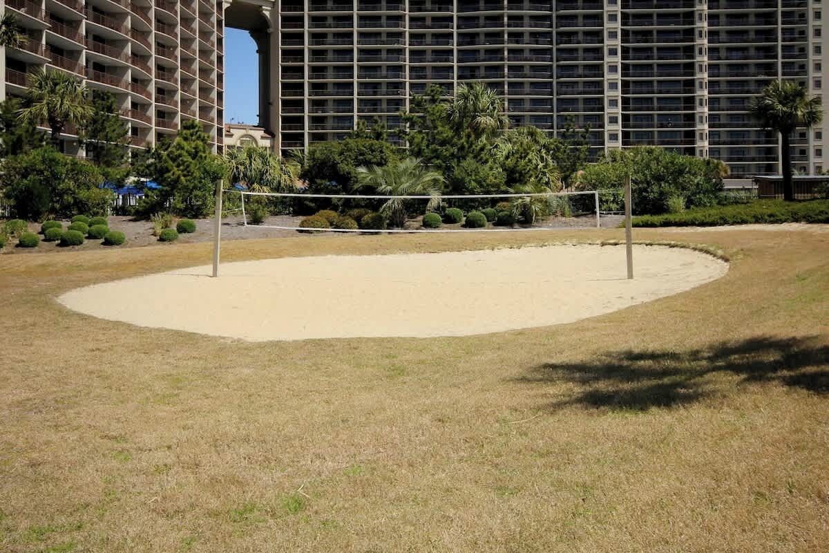 Ein Sandvolleyballfeld mit Netz, umgeben von einer Rasenfläche und Bäumen, mit einem großen Gebäude im Hintergrund.