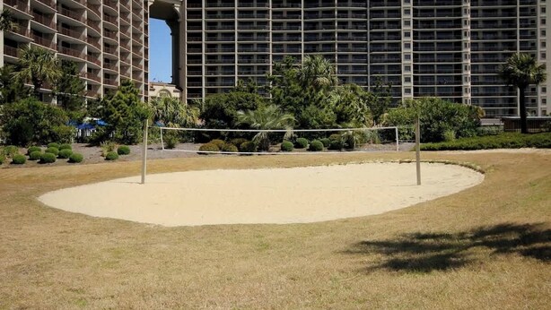 A sandy volleyball court with a net, surrounded by a grassy area and trees, with a large building in the background. - Ocean Creek Lodge IV - 2287 (Myrtle Beach)
