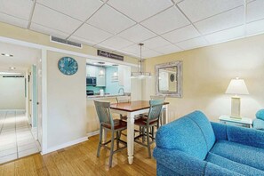 A room with a dining table, chairs, a blue sofa, a clock, and a kitchen area. - Ocean Creek Lodge IV - 2287 (Myrtle Beach)