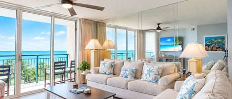 A spacious living room with a large sofa, a coffee table, and a TV. There are two ceiling fans, a large window with a view of the ocean, and a balcony with chairs.