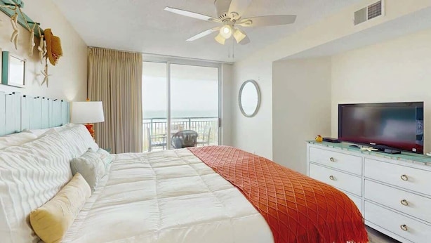 A hotel room with a bed, a television, a ceiling fan, and a view of the outdoors. - Windy Hill Dunes 204 (North Myrtle Beach)