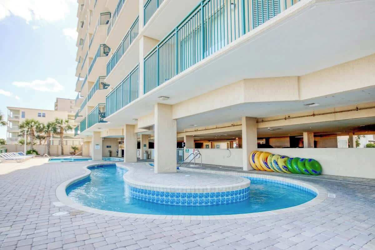 Zona de piscina de hotel con hidromasaje, sillas de descanso y un edificio con balcones.
