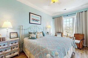 Une chambre à coucher comprenant un lit, des tables de chevet, une chaise, une fenêtre avec des rideaux et un ventilateur de plafond.