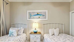 A bedroom with two beds, a nightstand with a lamp, and a wall-mounted painting of a fish. - Marsh Hawk 3D at Litchfield by the Sea (Pawleys Island)