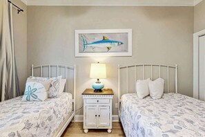 A bedroom with two beds, a nightstand with a lamp, and a wall-mounted painting of a fish. - Marsh Hawk 3D at Litchfield by the Sea (Pawleys Island)