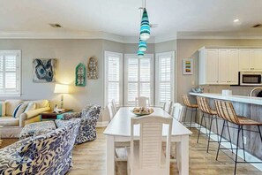 A dining area with a white table and chairs, a kitchen with white cabinets, and a hanging light fixture. - Marsh Hawk 3D at Litchfield by the Sea (Pawleys Island)