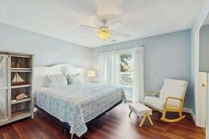 A bedroom with a bed, a rocking chair, a nightstand, a lamp, and a window with curtains. - Livin' the Dream at 26th Ave N (North Myrtle Beach)