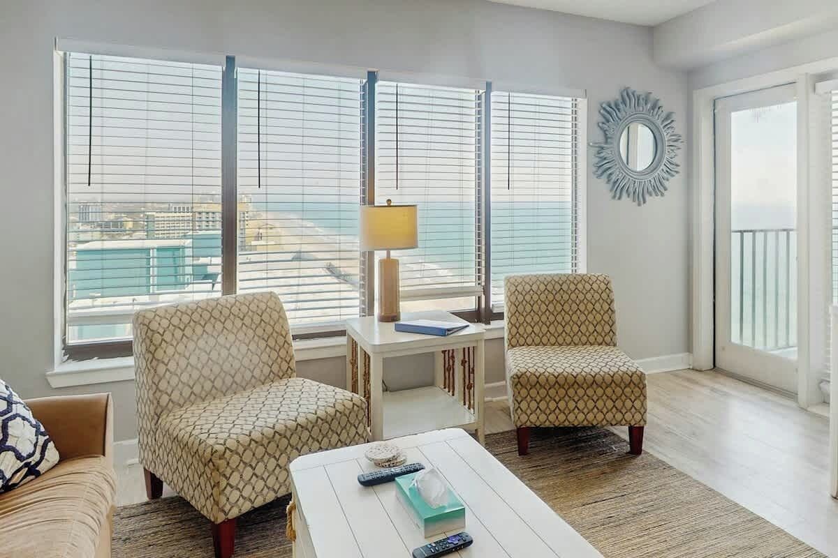 A living room with two patterned armchairs, a white coffee table with remote controls, a lamp, and a view of the city through large windows.