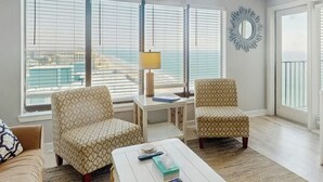 Uma sala de estar com dois poltronas estampadas, uma mesa de centro branca com controles remotos, um abajur e vista da cidade por grandes janelas.