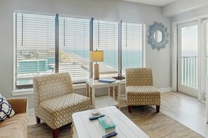 Ein Wohnzimmer mit zwei gemusterten Sesseln, einem weißen Couchtisch mit Fernbedienungen, einer Lampe und Blick auf die Stadt durch große Fenster.