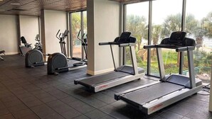 Et fitnesscenter med løbebånd, crosstrainere og en motionscykel, store vinduer med udsigt til palmer.