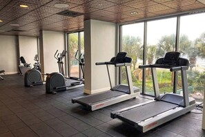 Ein Fitnessraum mit Laufbändern, Crosstrainern und einem Ergometer, große Fenster mit Blick auf Palmen.